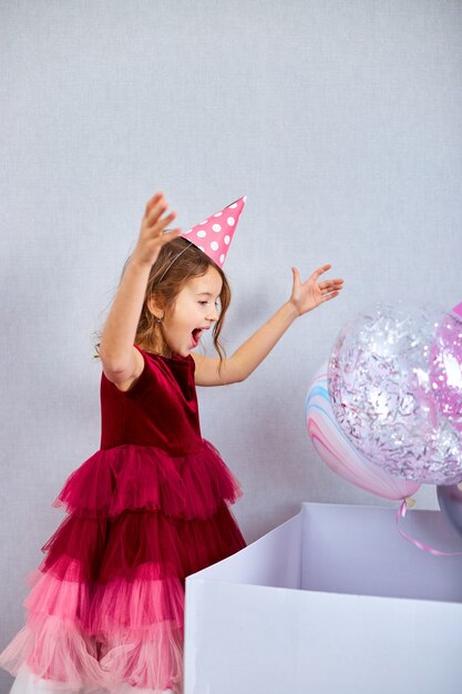 Menina alegre de vestido rosa e chapéu abre uma grande caixa de presente com balões