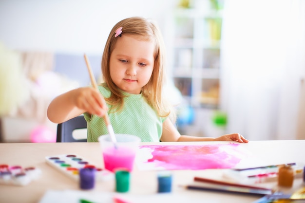 Menina alegre desenha guache em cores diferentes em uma folha de papel branca.