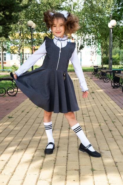 Menina alegre e engraçada com um sorriso desdentado em um uniforme escolar com laços brancos no pátio da escola De volta à escola, 1º de setembro Um aluno feliz Educação primária Classe primária Retrato de um estudante