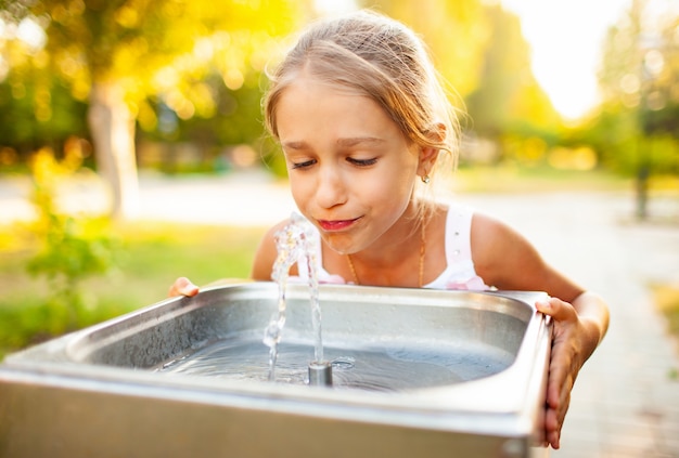 Menina alegre e maravilhosa bebe água fresca de uma pequena fonte em um parque ensolarado e quente de verão em férias há muito aguardadas