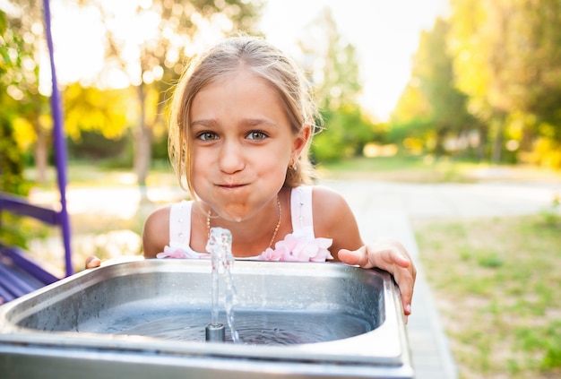 Menina alegre e maravilhosa bebe água fresca de uma pequena fonte em um parque ensolarado e quente de verão em férias há muito aguardadas