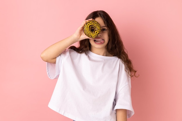 Menina alegre em pé se divertindo segurando e cobrindo os olhos com rosquinha e mostrando a língua