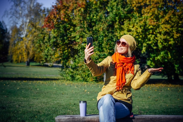 Menina alegre em roupas brilhantes e óculos de sol, sentado no banco do parque e tomando selfie no smartphone contra árvores de outono. mulher atraente faz gestos com as mãos no fundo da natureza. copie o espaço.