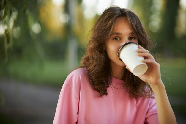 Menina alegre em uma camiseta rosa caminha no parque e bebe café para viagem de um copo de papel.