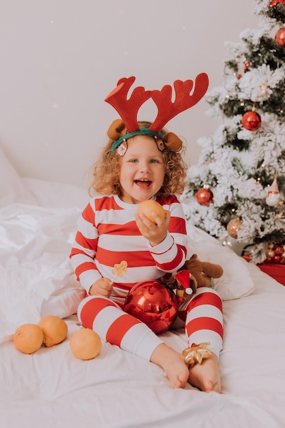 Menina alegre encaracolada de pijama de natal come pirulito e tangerinas sentada na cama criança é