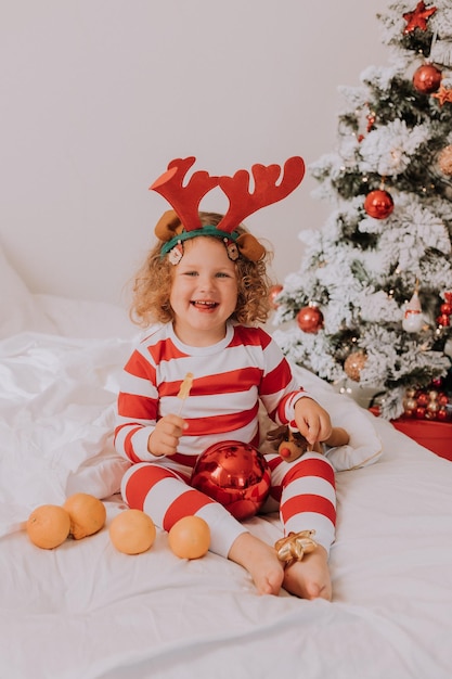 Menina alegre encaracolada de pijama de Natal come pirulito e tangerinas sentada na cama. criança está curtindo o Natal. estilo de vida. espaço para texto. Foto de alta qualidade