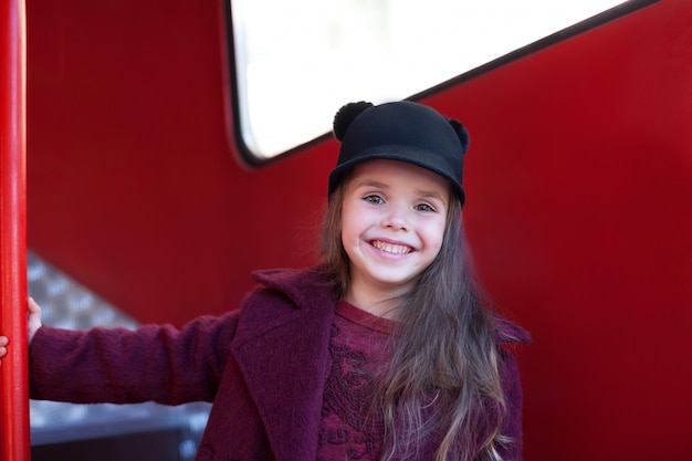 Menina alegre perto do ônibus vermelho inglês em um lindo casaco e chapéu. Menina alegre perto do ônibus vermelho inglês em um lindo casaco e um chapéu. Jornada da criança. Ônibus escolar. Ônibus vermelho de Londres.