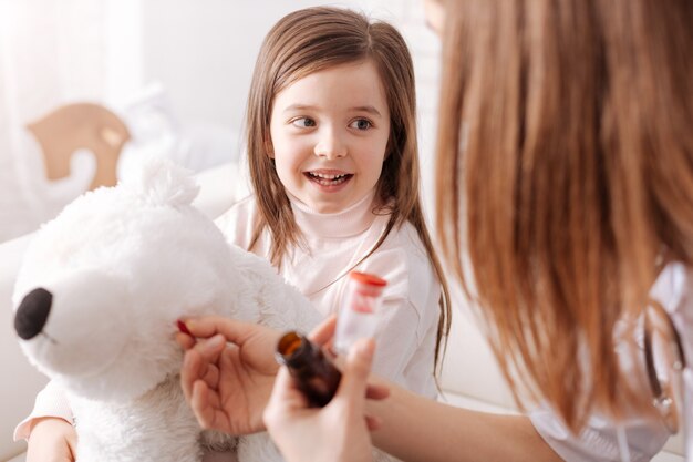 Menina alegre segurando seu brinquedo fofo enquanto a mãe dela finge ser médica e dá comprimidos