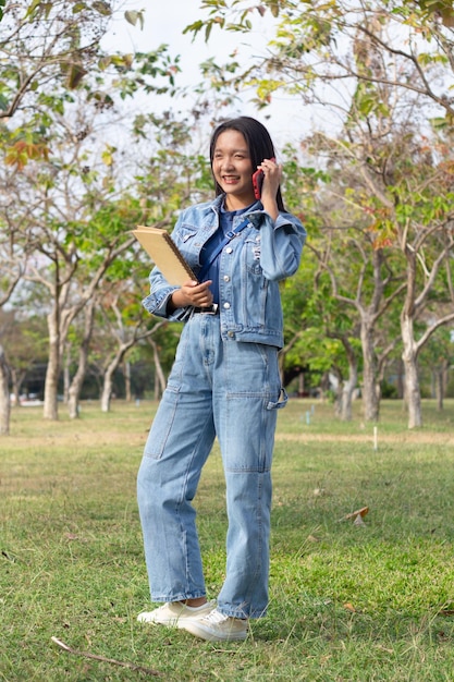 Menina alegre usando um telefone celular e segurando um livro parado no parque usa jaqueta e jeans conceito de estilo de vida