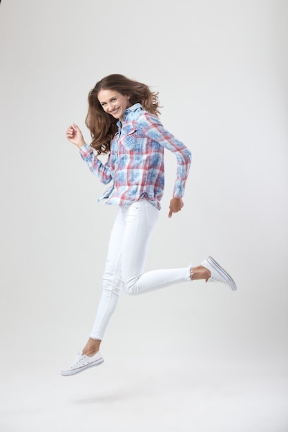 Menina alegre vestida com uma camisa xadrez e jeans branco salta sobre o fundo branco