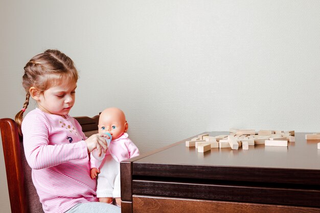 Menina alimentando uma boneca enquanto está sentada à mesa