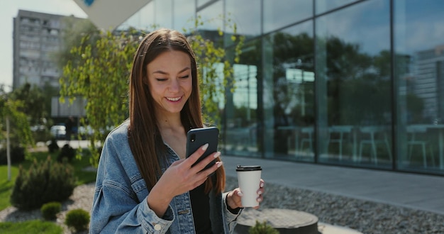 Menina andando com smartphone e xícara de café Linda mulher usando telefone com café para viagem na rua