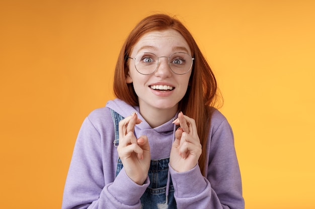 Menina animada assistindo resultados dedos cruzados boa sorte torcendo time favorito quer ganhar o sonho tornado realidade emocionado boca aberta olhar fixo câmera esperançoso suplicando desejo cumprido, fundo laranja.