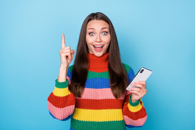 Menina animada com retrato usando smartphone, pensamentos, ter uma ideia incrível de que mensagens de texto digitando rede social levantar o dedo indicador usar suéter estilo arco-íris isolado fundo de cor azul