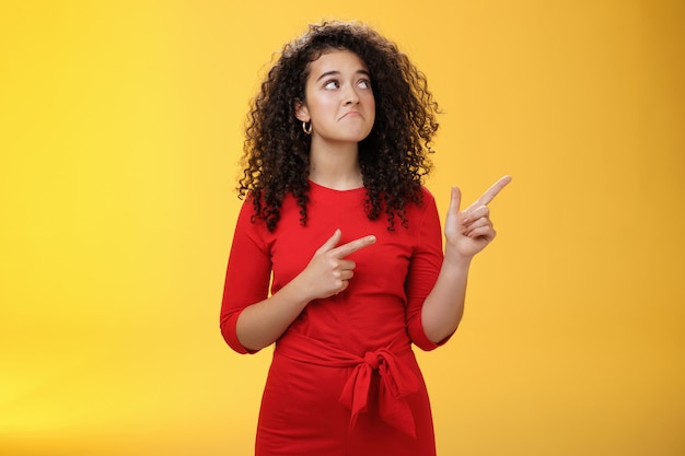 Menina apontando e olhando para o canto superior direito com pesar e tristeza perdendo uma boa chance ou oportunidade ficando chateada e triste, puxando um sorriso sombrio com os lábios franzidos em pé sobre a parede amarela.