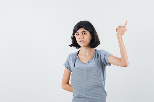 Menina apontando para cima em t-shirt, avental e parecendo perplexa. vista frontal.