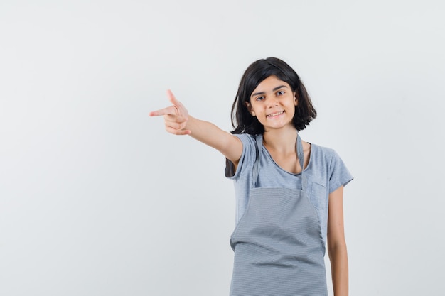 Menina apontando para longe em t-shirt, avental e parecendo confiante. vista frontal.