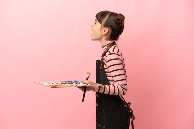 Menina artista segurando uma paleta isolada na rosa rindo na posição lateral