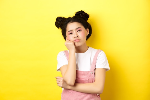 Menina asiática adolescente triste e entediada sozinha, olhando para a câmera indiferente, olhando com tédio, amarelo.
