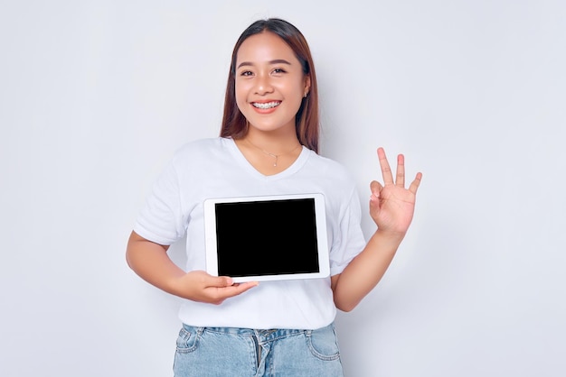 Menina asiática alegre em camiseta branca mostrando tablet digital com tela vazia e sinal de ok isolado no anúncio de fundo branco para um aplicativo ou site