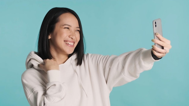 Menina asiática alegre vestindo capuz fazendo uma chamada de vídeo usando seu smartphone conversando alegremente com o melhor amigo sobre fundo azul Tecnologia moderna