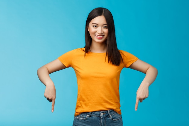 Menina asiática autoconfiante alegre corte de cabelo escuro usar camiseta amarela de verão apresentar promoção, apontando as mãos para baixo, indicar anúncio de fundo sorrindo encantado, compartilhar link bom fundo azul