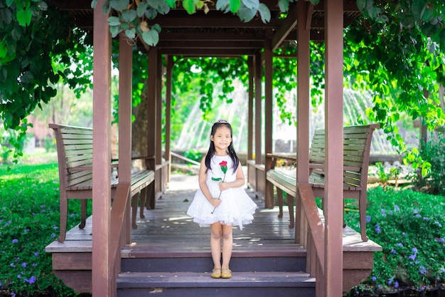 Foto menina asiática bonitinha em um vestido branco segurando uma rosa vermelha em pé no pavilhão