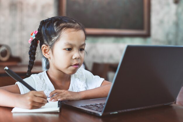 Menina asiática bonito da criança que usa o portátil e escrevendo em seu caderno no café