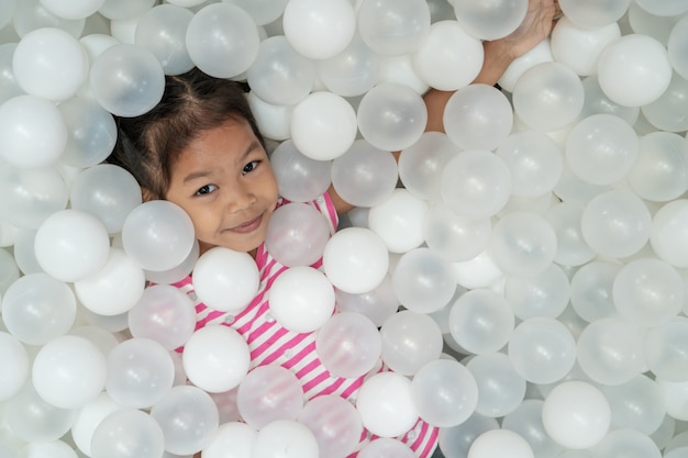 Menina asiática bonito feliz da criança que tem o divertimento a jogar com as bolas plásticas brancas no campo de jogos