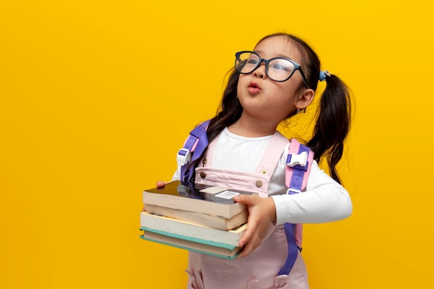 Menina asiática com óculos e mochila segurando livros em fundo isolado amarelo