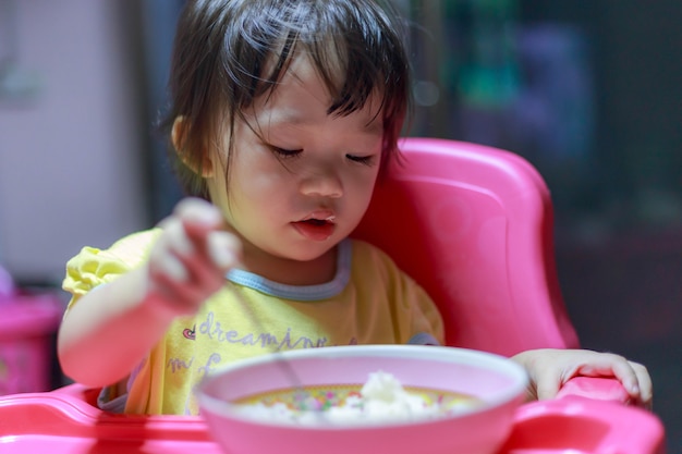 Menina asiática comendo salsichas fritas na tigela de alumínio perto da janela em casa