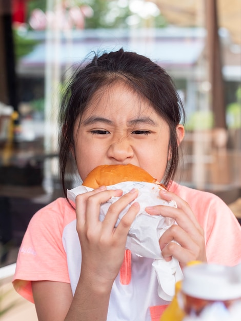 Menina asiática comendo um hambúrguer. As crianças asiáticas comem queijo de frango Hambúrguer.