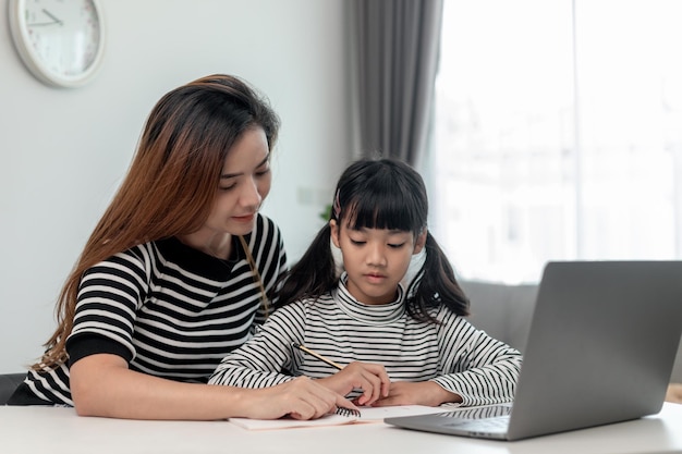 Menina asiática criança aprendendo aula on-line em casa com a mãe criança pré-escolar usa laptop para fazer lição de casa do professor da escola pela internet remota digital com apoio da mãe