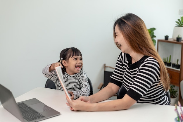 Menina asiática criança aprendendo aula on-line em casa com a mãe Criança pré-escolar usa laptop para fazer lição de casa do professor da escola pela internet remota digital com apoio da mãe