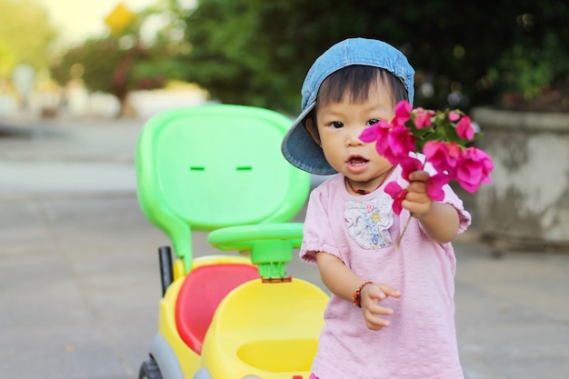 Menina asiática da criança do bebê que guarda flores cor-de-rosa em sua mão e que joga um brinquedo do carro no campo de jogos.