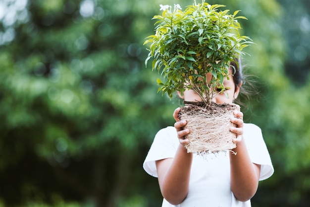 Menina asiática da criança que guarda a árvore nova para prepara o plantio no solo como salvar o conceito do mundo