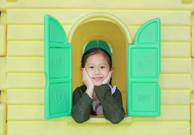 Menina asiática da criança que joga com o teatro do brinquedo da janela no campo de jogos.