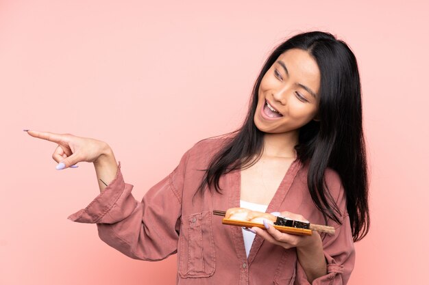 Menina asiática de adolescente comendo sushi na parede rosa, apontando o dedo para o lado