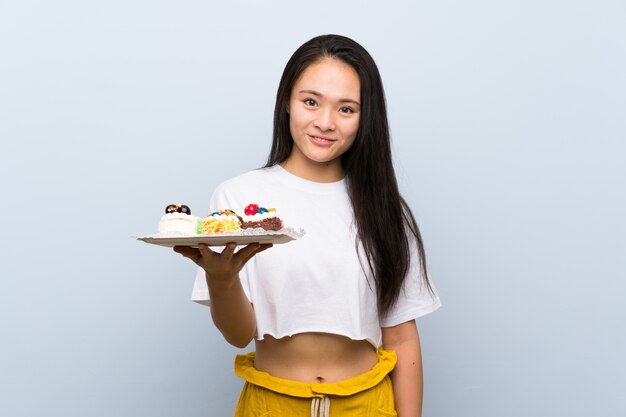 Menina asiática de adolescente segurando muitos mini bolos diferentes, sorrindo muito