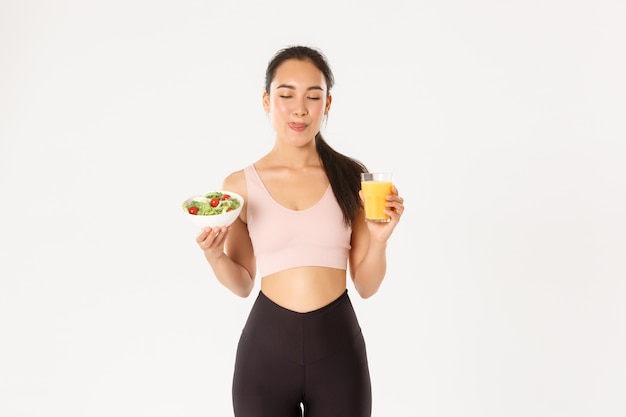 Menina asiática de aptidão fofa sorridente no activewear, lambendo os lábios, satisfeita com uma deliciosa salada saudável e suco de laranja antes do treino no ginásio.