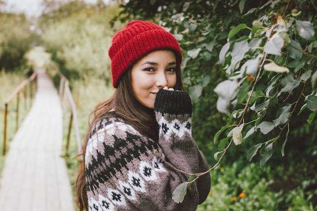 Menina asiática de cabelo comprido despreocupado bonito de chapéu vermelho e camisola nórdica de malha no parque natural de outono, estilo de vida de aventura de viagem