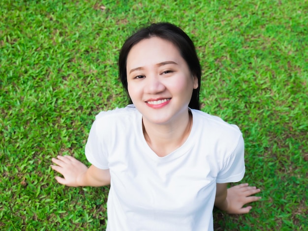 Menina asiática de camisa branca saia para relaxar e aproveitar a luz do sol quente à noite Em um feliz dia de descanso do trabalho Lá fora sentado no gramado verde olhando para a luz e sorrindo no parque
