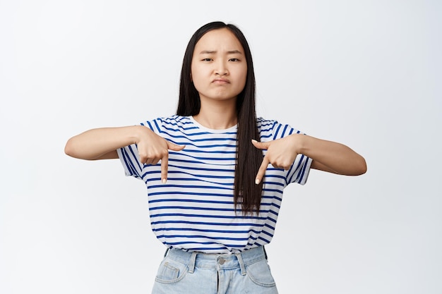 Menina asiática desapontada, apontando os dedos para baixo, franzindo a testa, chateada e mostrando um produto de aversão ruim contra o fundo branco