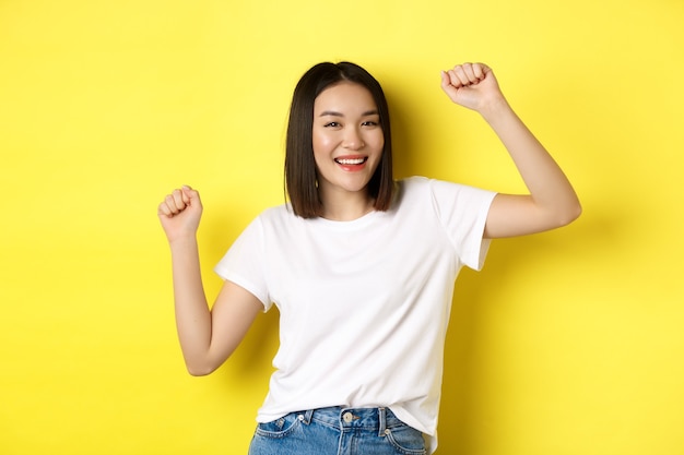 Menina asiática despreocupada dançando e se divertindo, posando com uma camiseta branca contra um fundo amarelo