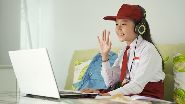 Menina asiática do ensino fundamental aprendendo saudação online na tela do laptop em casa