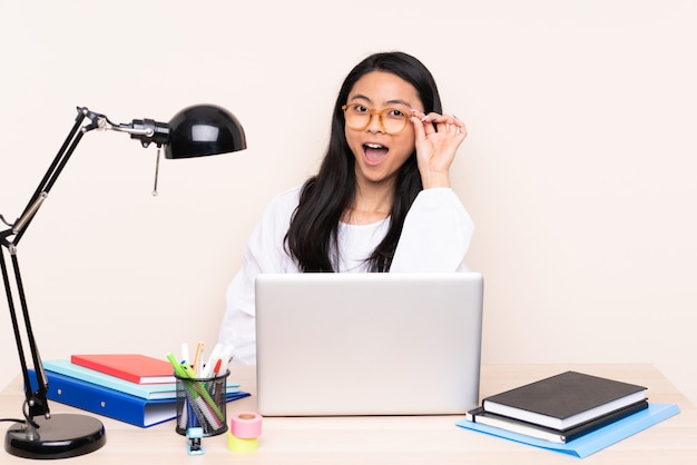 Menina asiática do estudante em um local de trabalho com um laptop isolado na parede bege com óculos e surpreso