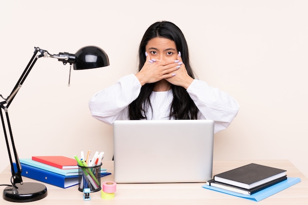 Menina asiática do estudante em um local de trabalho com um laptop isolado no bege