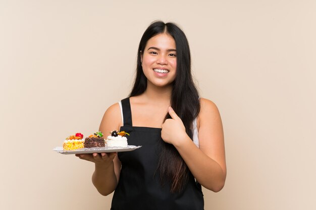 Menina asiática do jovem adolescente segurando muitos mini bolos diferentes sobre parede isolada com expressão facial de surpresa