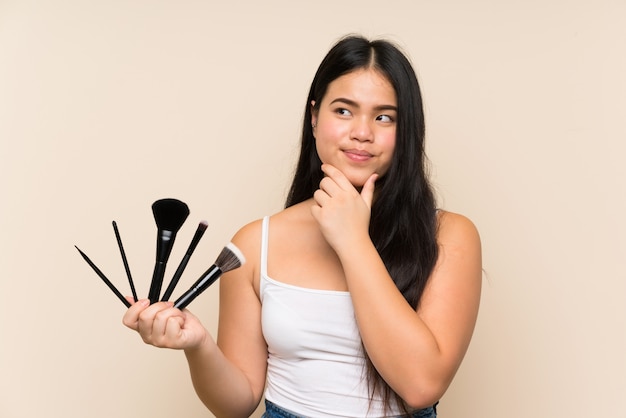 Menina asiática do jovem adolescente segurando um monte de pincel de maquiagem, pensando em uma idéia