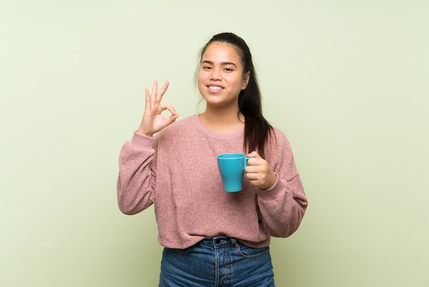 Menina asiática do jovem adolescente sobre parede verde isolada, segurando a xícara de café quente